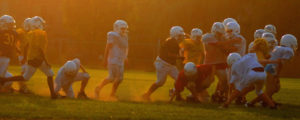 A group of young boys playing football wearing helmets, pads, and jerseys on a green field as the sun sets