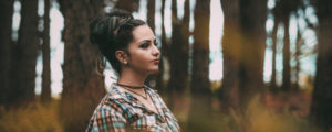 Brunette woman standing in a forest while wearing a plaid shirt looks away anxiously as she thinks about the dentist
